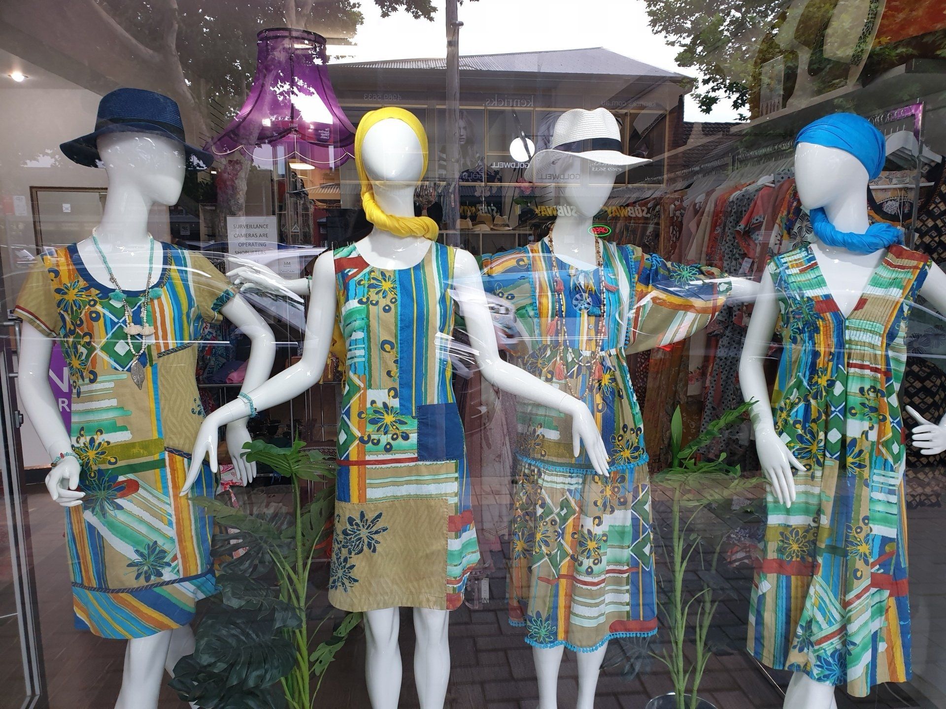 Four mannequins in a shop window display wearing colorful dresses and hats.