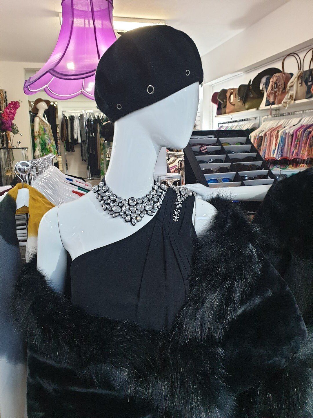 Mannequin in black dress, fur wrap, hat, and beaded necklace, inside a clothing store.