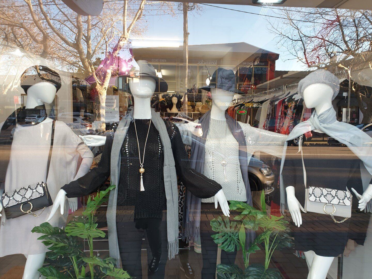 Four mannequins in a store window display, wearing hats and accessories. A small plant is in front of them.