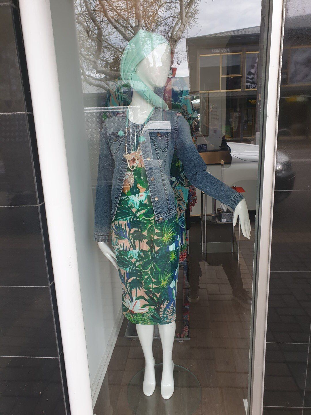 Mannequin in window display wearing a floral dress, denim jacket, and headscarf.