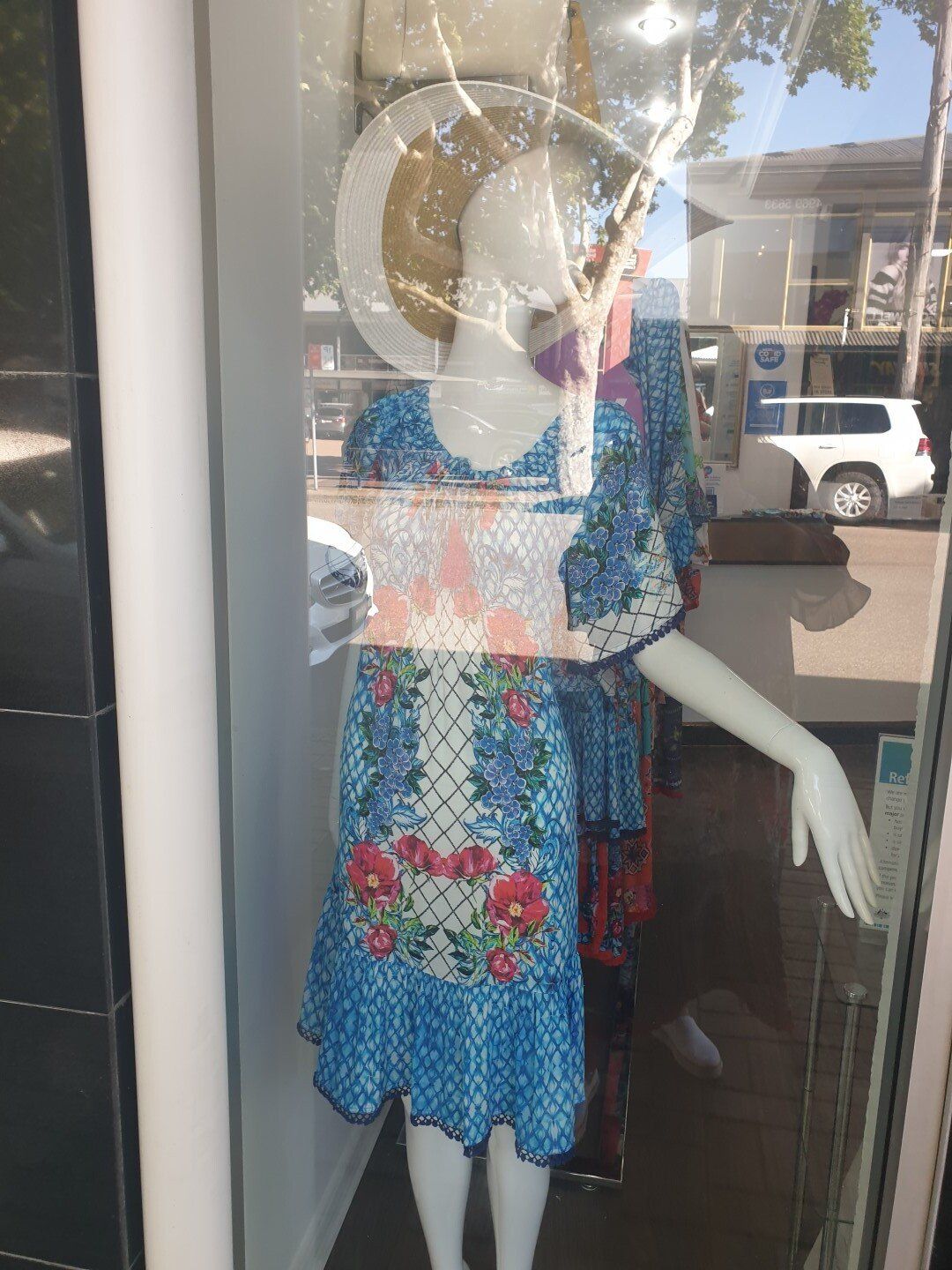 Mannequin in a window display wearing a blue floral dress and a straw hat, with a city street background.