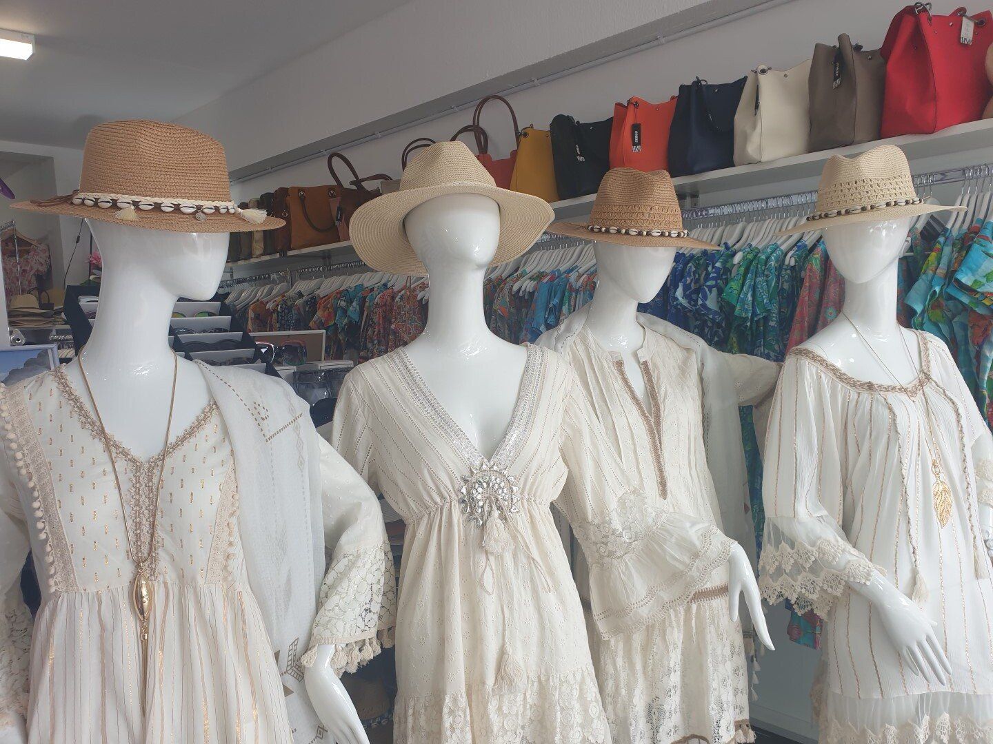 Four mannequins wearing white dresses and straw hats in a shop, with colorful bags on a shelf behind them.