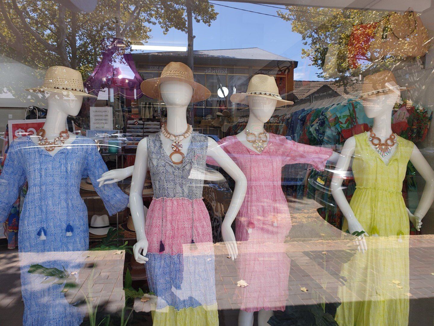 Four mannequins in a shop window wearing summer dresses, hats, and necklaces.