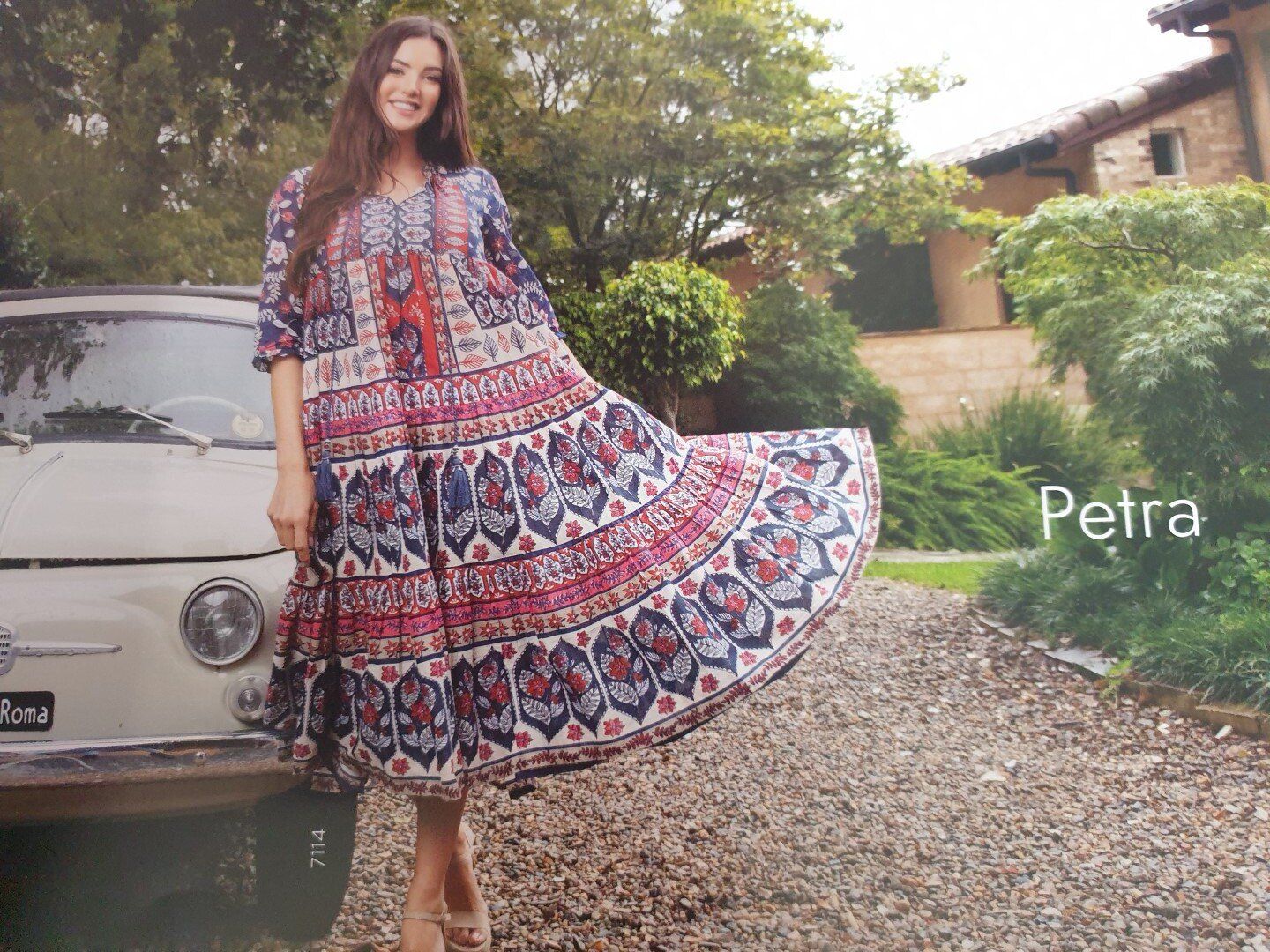 Woman in patterned dress smiles, standing next to vintage car, gravel driveway, green foliage, sunny day.