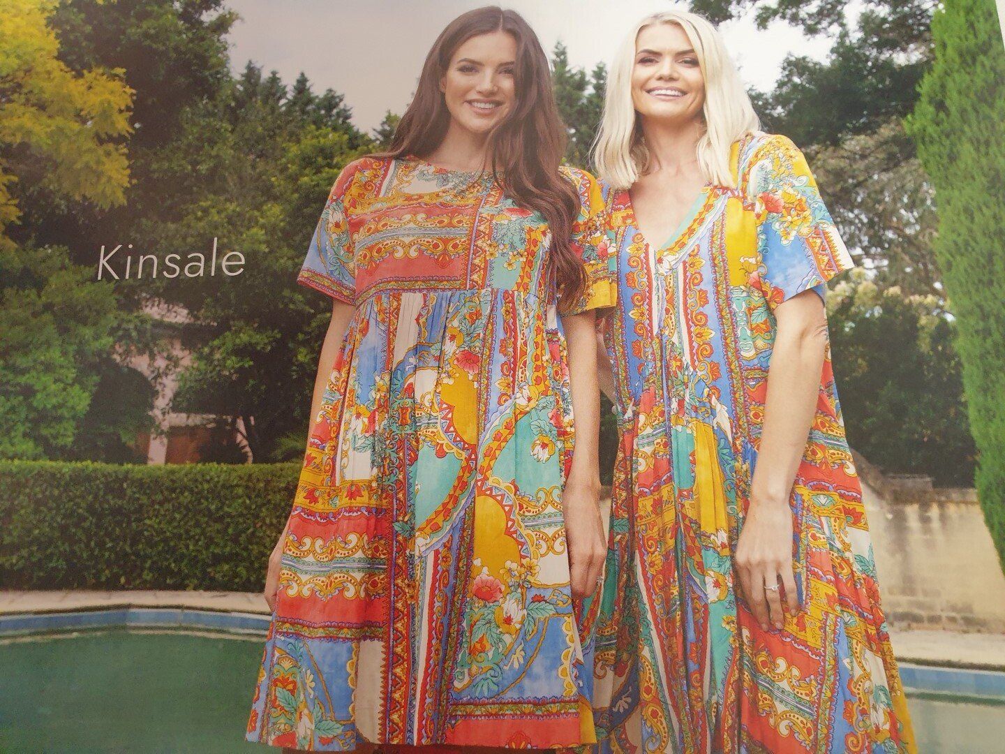 Two women wearing colorful patterned dresses, standing by a pool in a sunny outdoor setting.