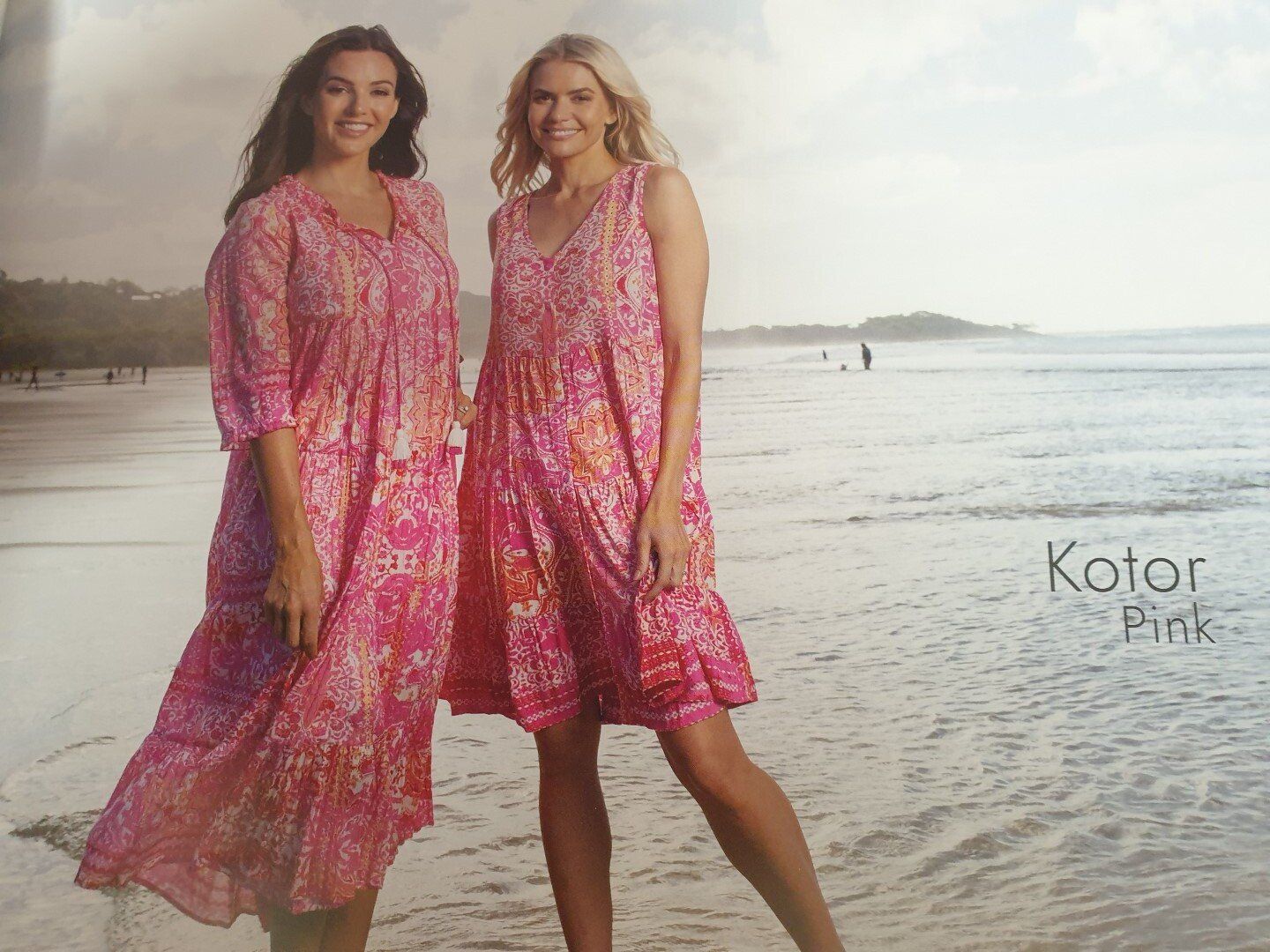 Two women in pink floral dresses on a beach. One smiles, the other poses with a leg forward. Ocean in the background.
