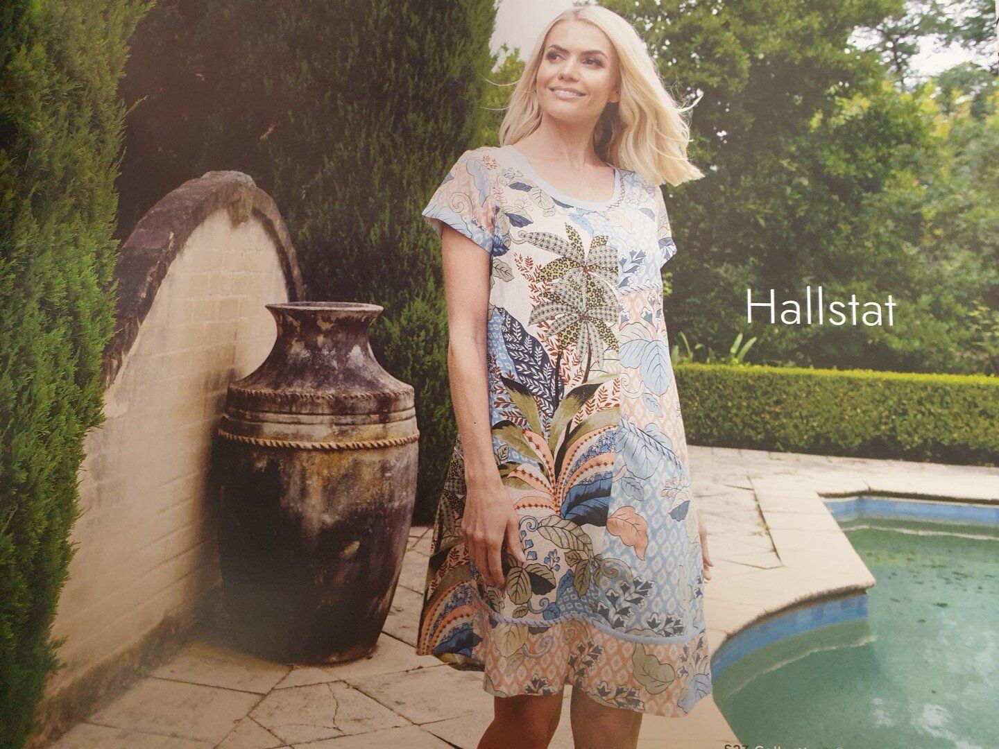 Woman in a floral dress smiles near a pool.  Hallstat text on right. Brown urn and green hedges are in the background.