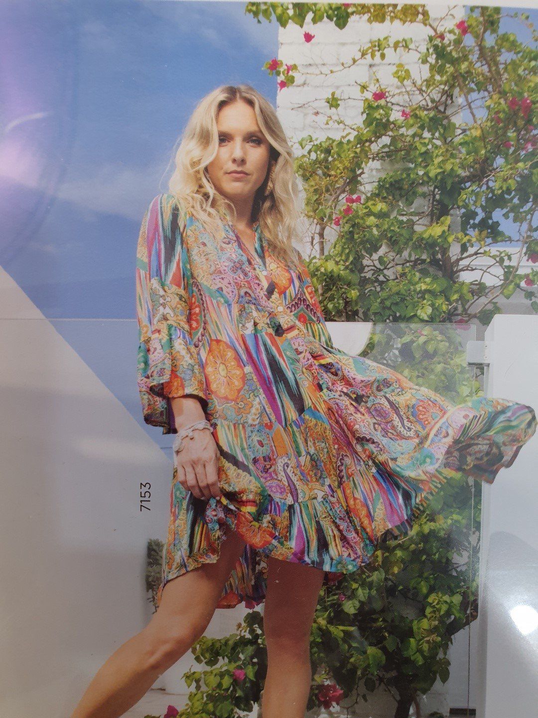 Blonde woman in colorful patterned dress, outside near white walls and green plants.