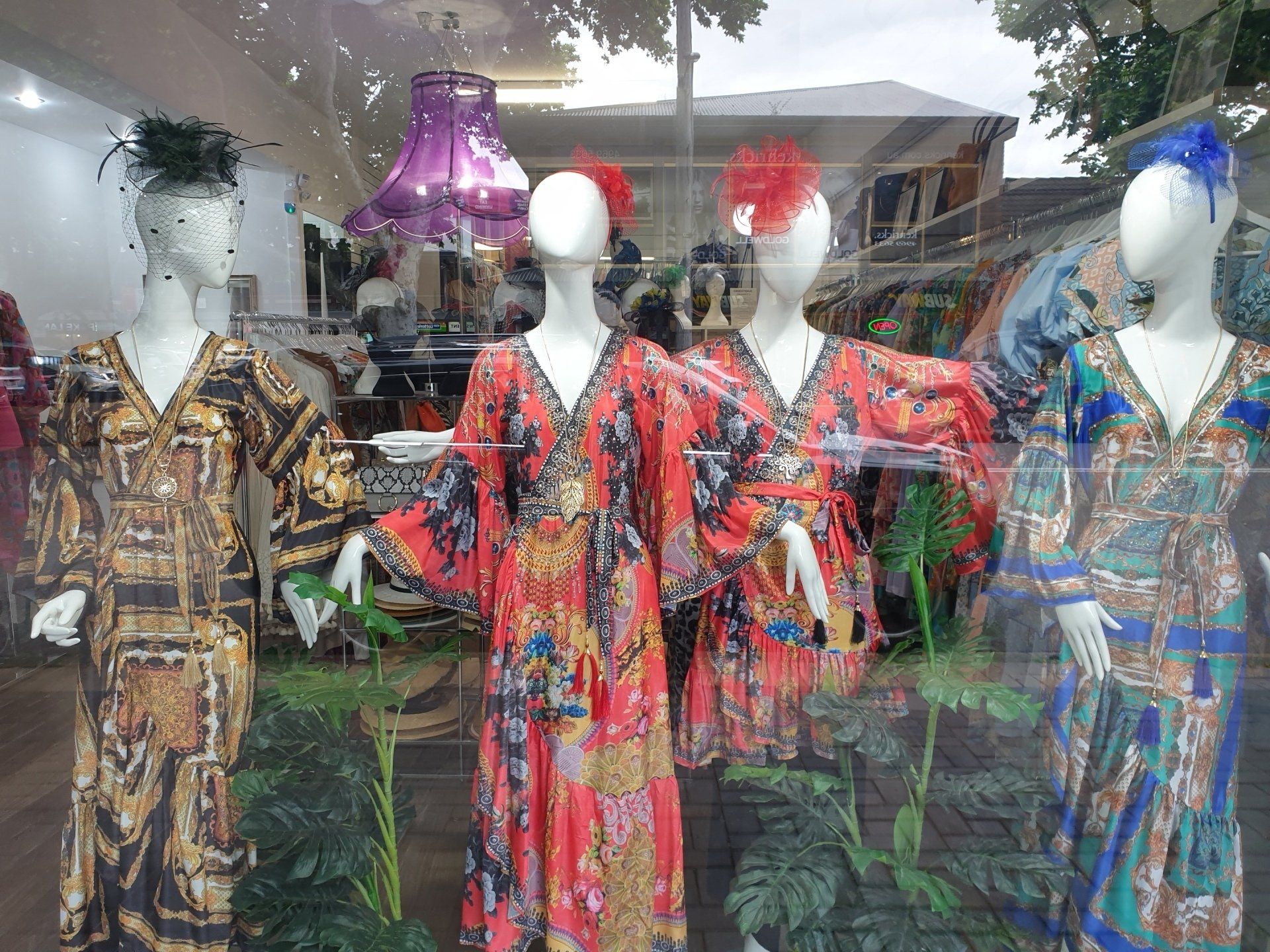 Mannequins in a shop window display colorful patterned dresses, various headpieces, and greenery.