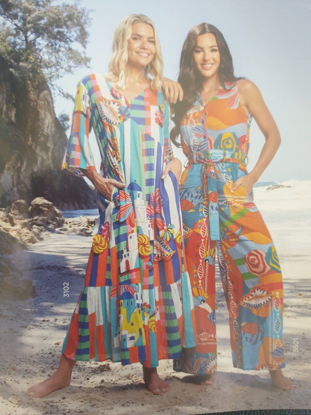 Two women on a beach wearing colorful patterned dresses and jumpsuit, smiling.
