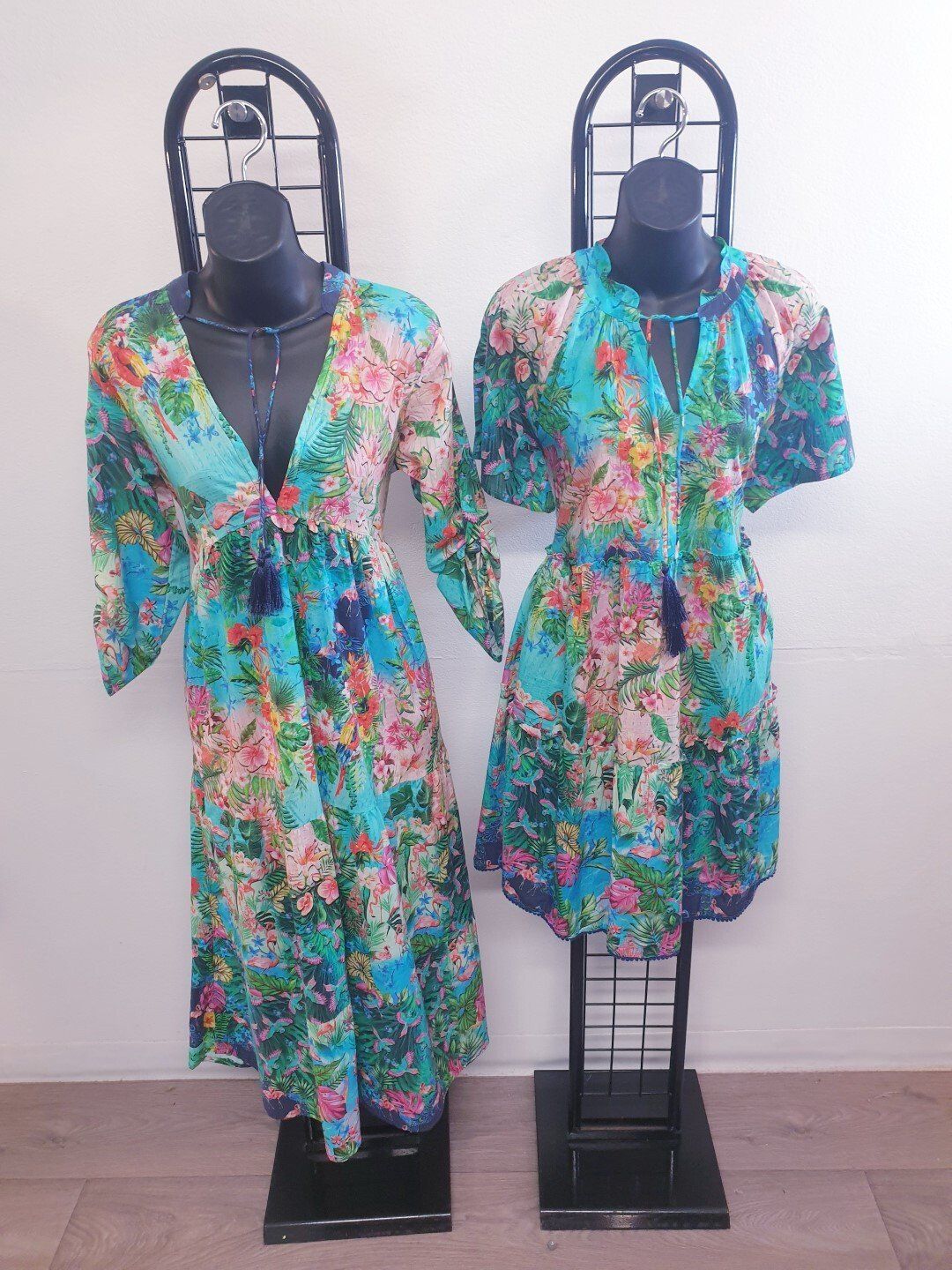 Two floral dresses on mannequins; one long-sleeved, one short-sleeved. Both are turquoise with pink and yellow flowers.