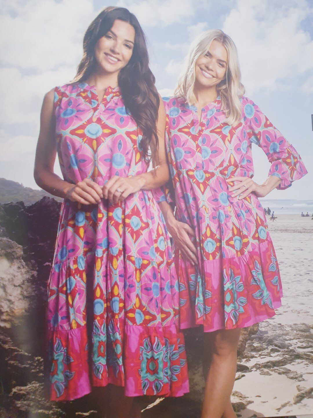 Two women smiling, wearing pink floral dresses, on a beach.