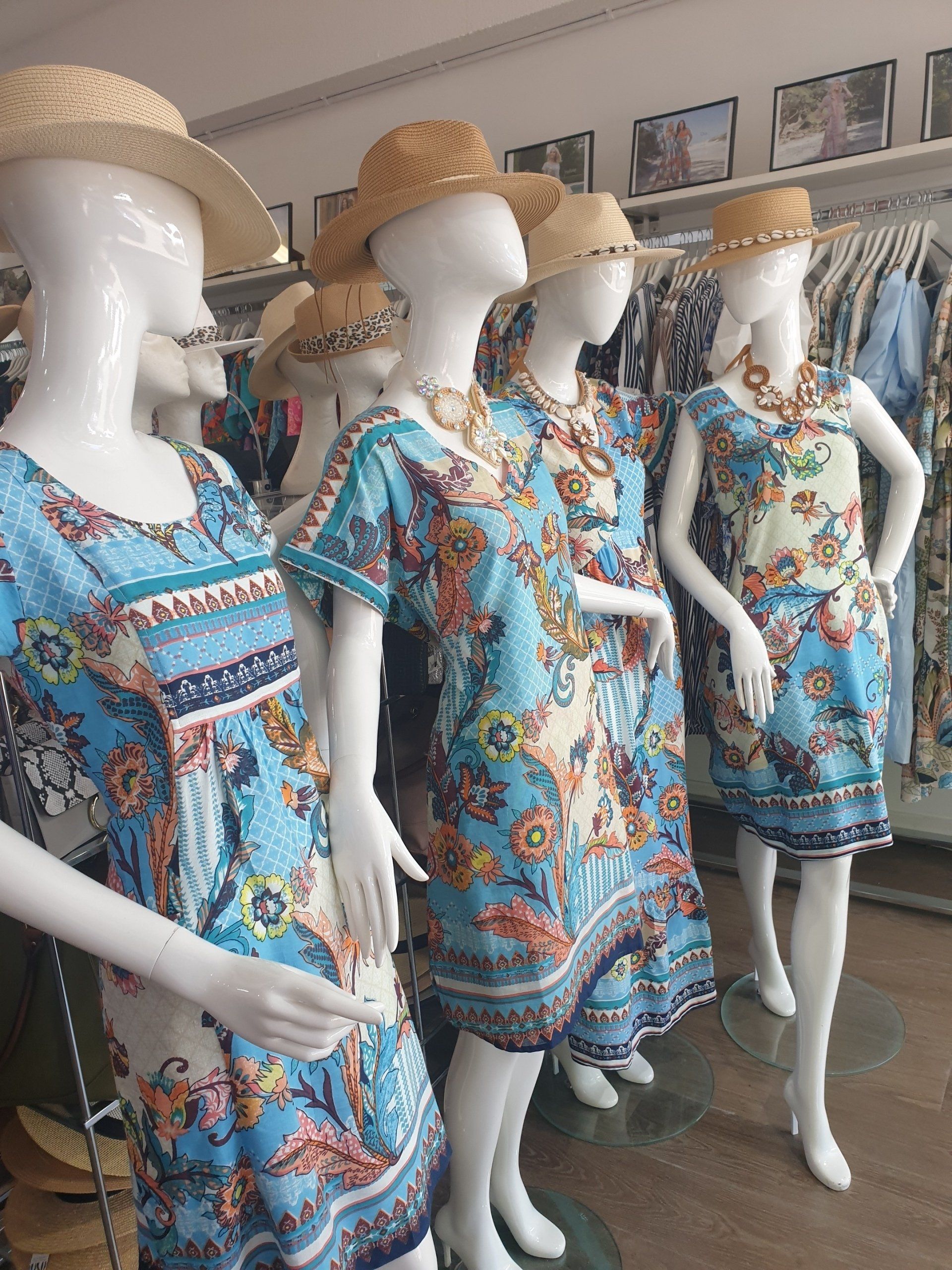 Mannequins wearing blue floral dresses, straw hats, and necklaces in a clothing store.