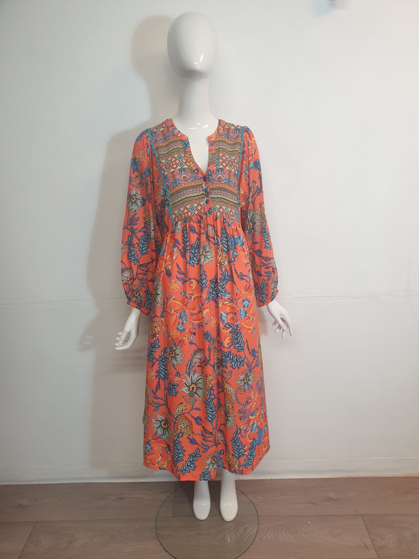 Mannequin in a long, orange floral dress with long sleeves, standing against a white wall.