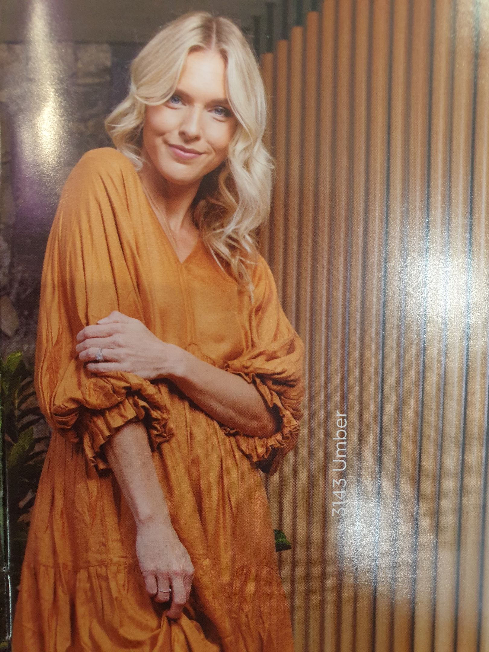 Blonde woman in a mustard-colored dress, smiling, leaning against a wood-paneled wall.