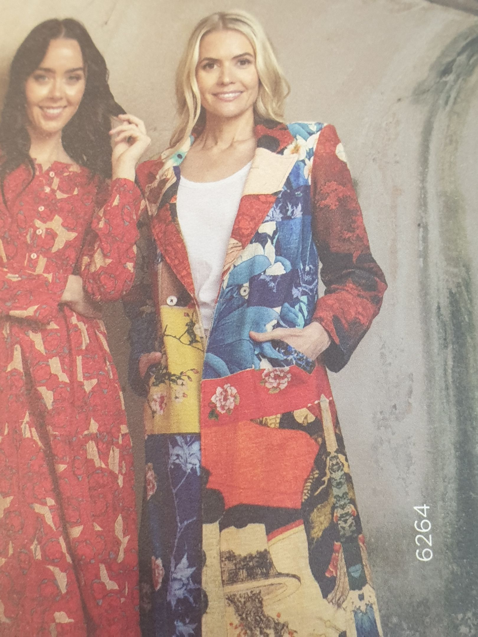 Woman in colorful patchwork coat; smiling, standing in front of wall.