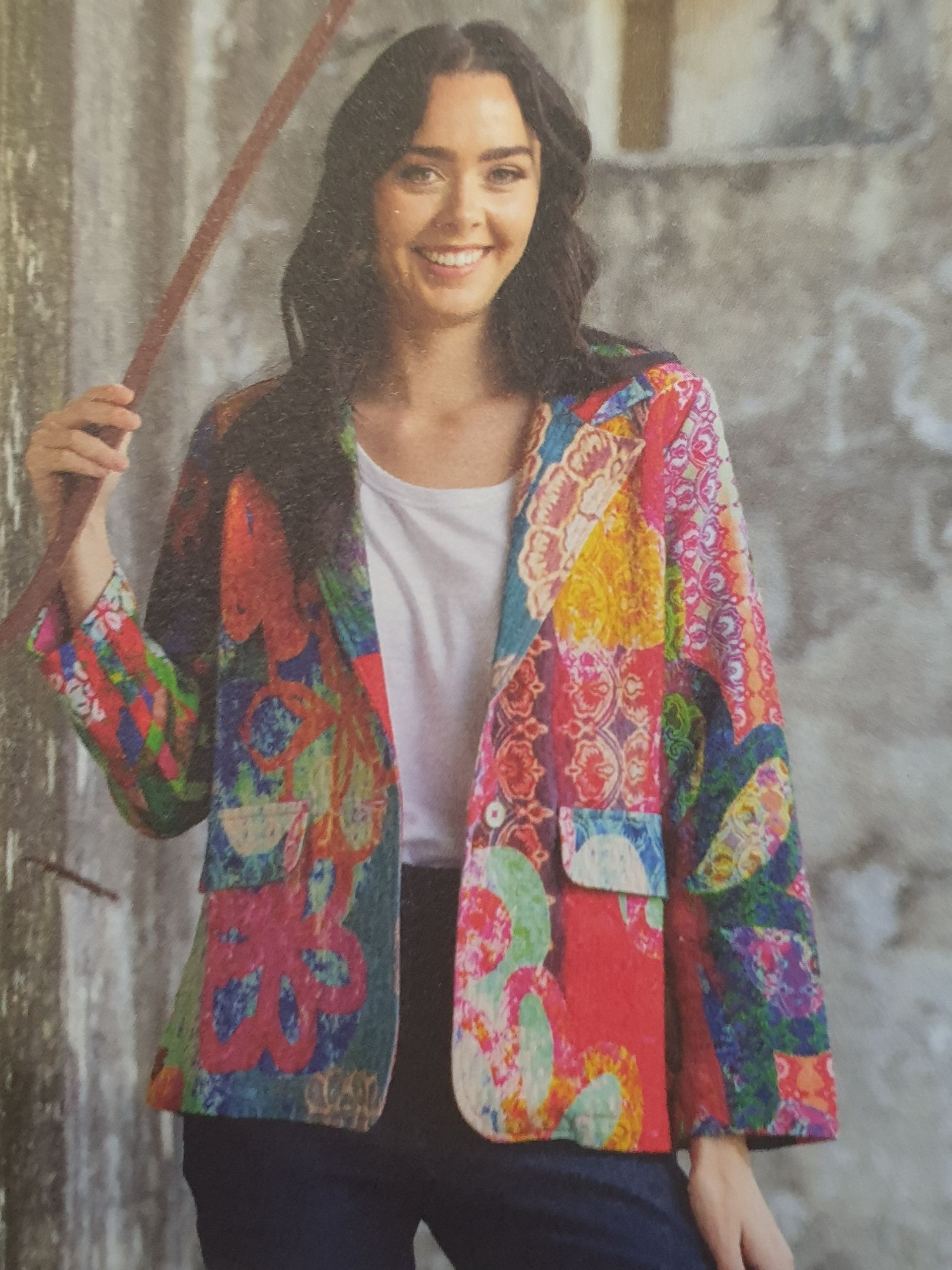 Woman in colorful patchwork blazer, smiling against a textured concrete backdrop.