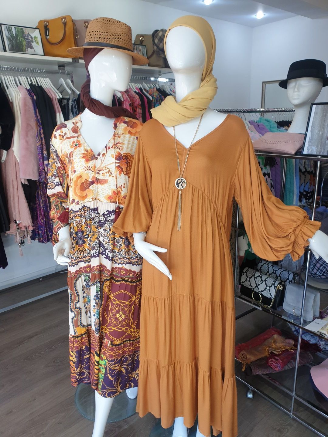 Two mannequins in a clothing store wearing dresses and scarves. Other clothes and hats on display.