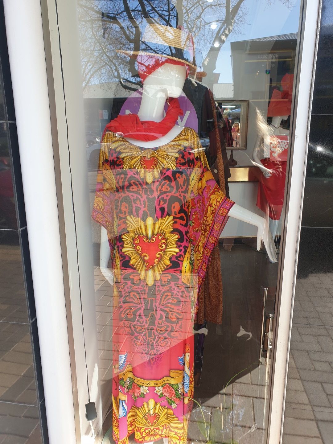 Mannequin in a shop window wearing a pink and gold patterned dress, hat, and red scarf.