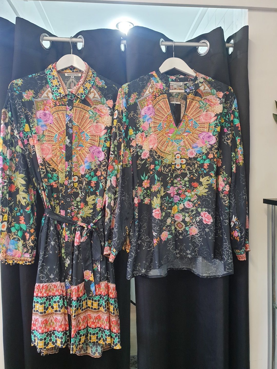 Two floral print shirts, one belted dress-style, hanging on black rods against a dark backdrop.