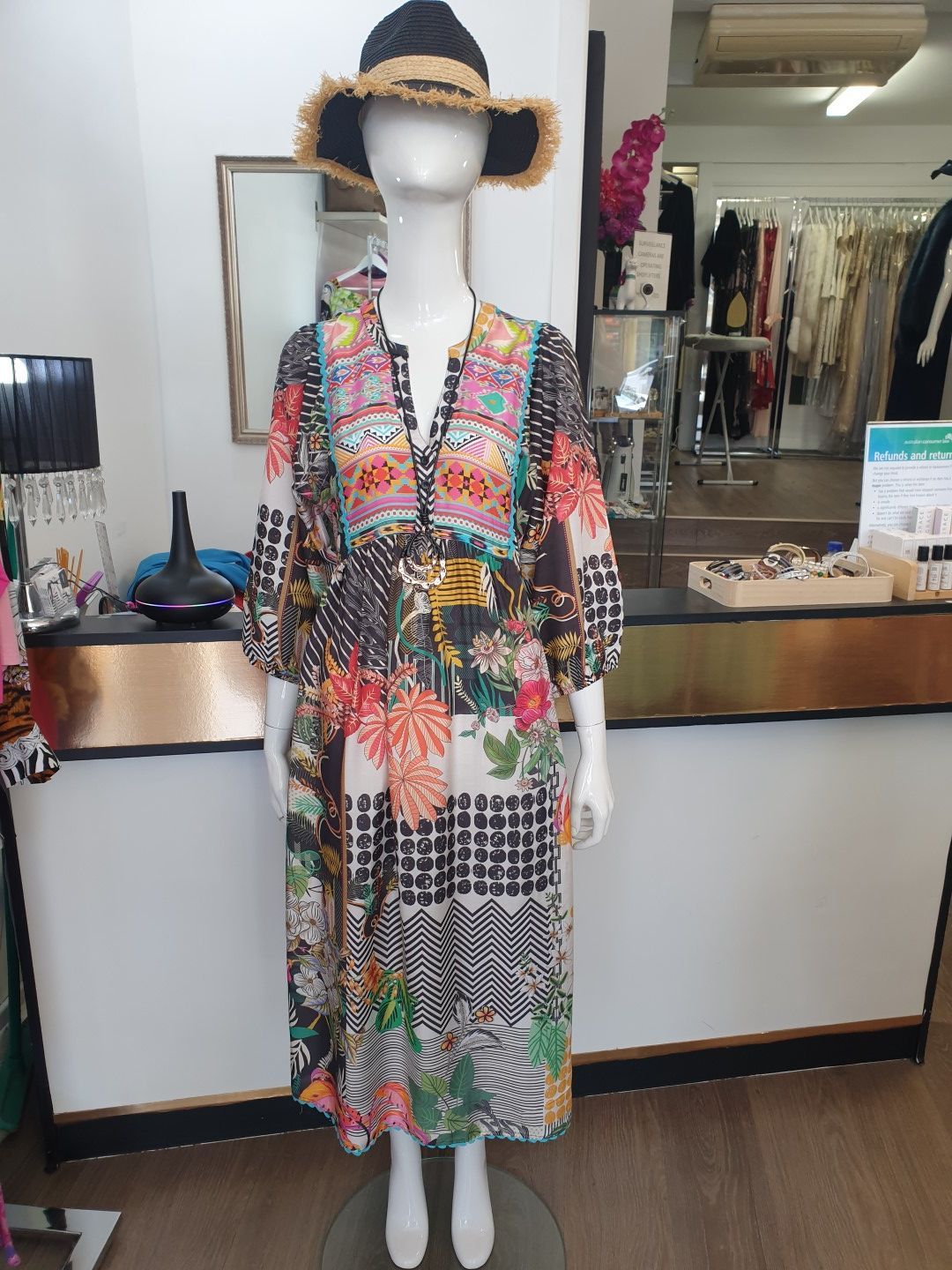 Mannequin in patterned dress and hat in a boutique, standing behind a counter.