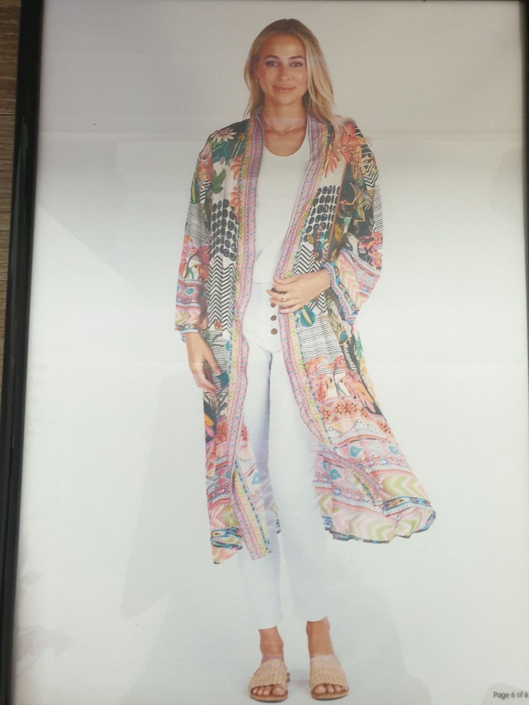 Woman in colorful floral duster coat, white pants, tan sandals, smiling.