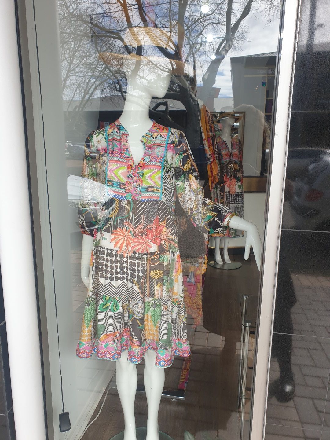 Mannequin in a shop window wearing a colorful patterned dress; outside trees reflect in the glass.
