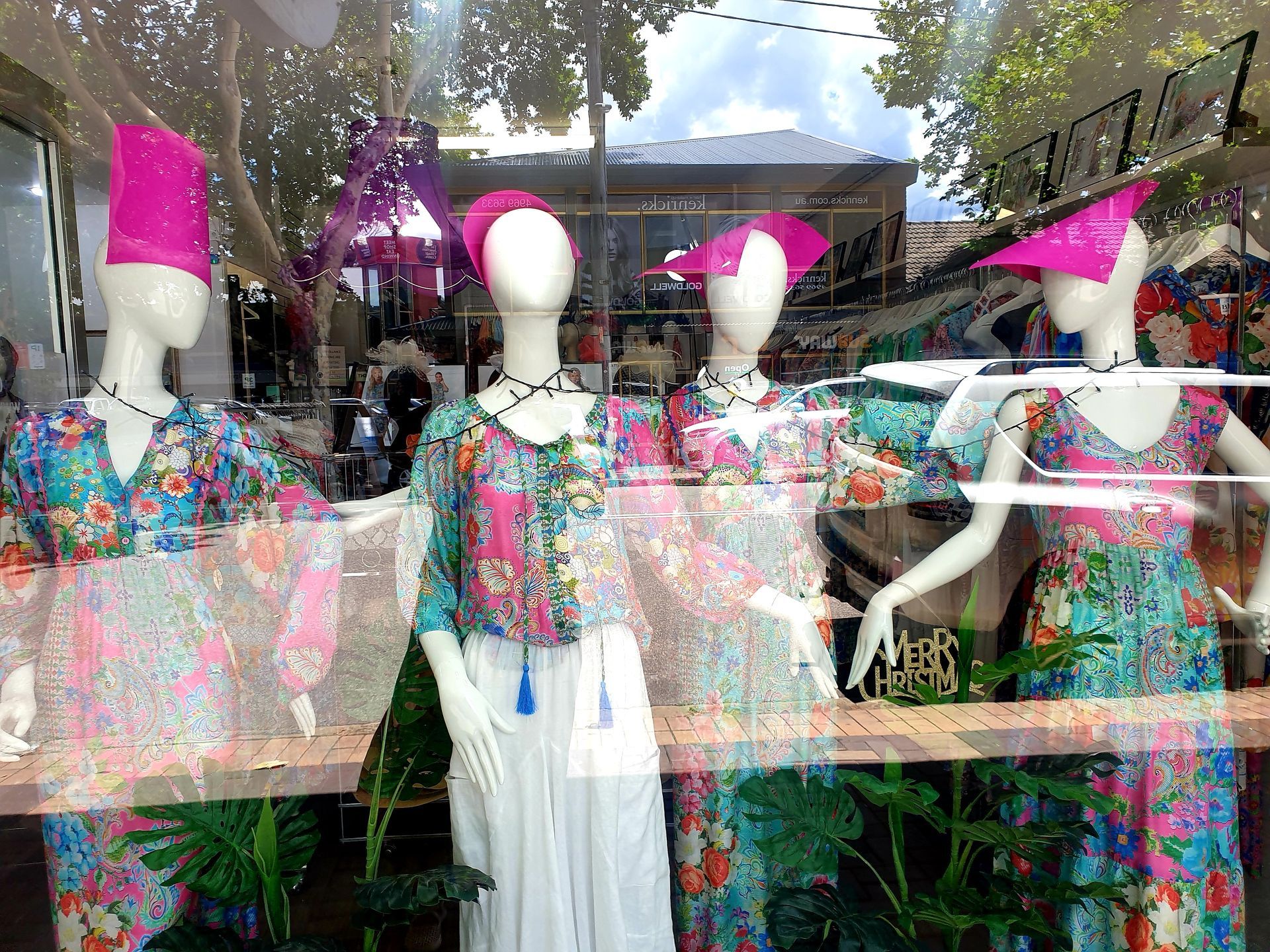 Mannequins in floral dresses and pink hats in a shop window, greenery visible.