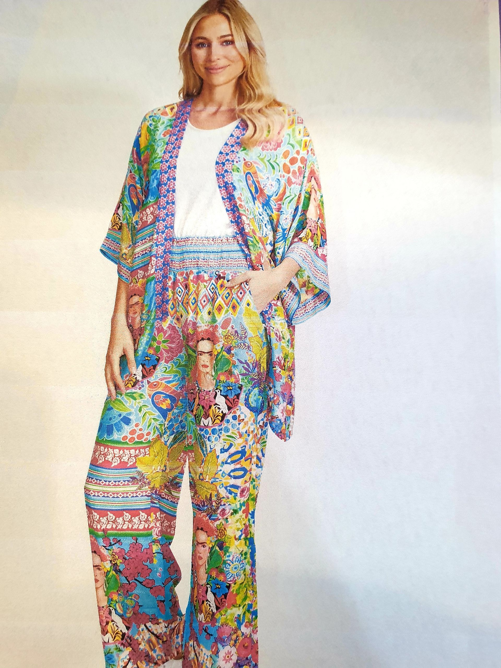 Woman wearing colorful floral kimono, pants, and white top, standing indoors with a neutral background.