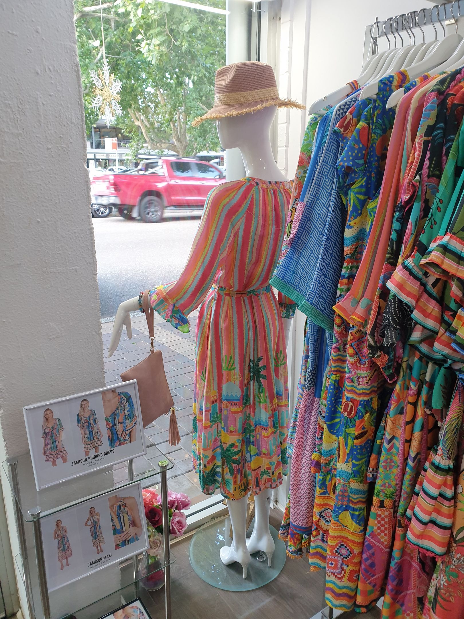 Mannequin in colorful dress, displayed in a boutique window next to a rack of similar dresses.