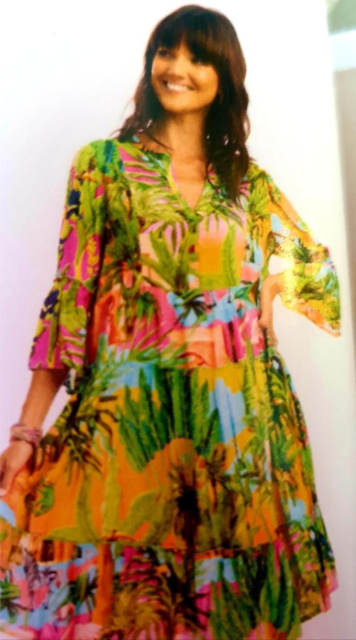 Woman in colorful tropical print dress smiles, standing outside, hands on hip.