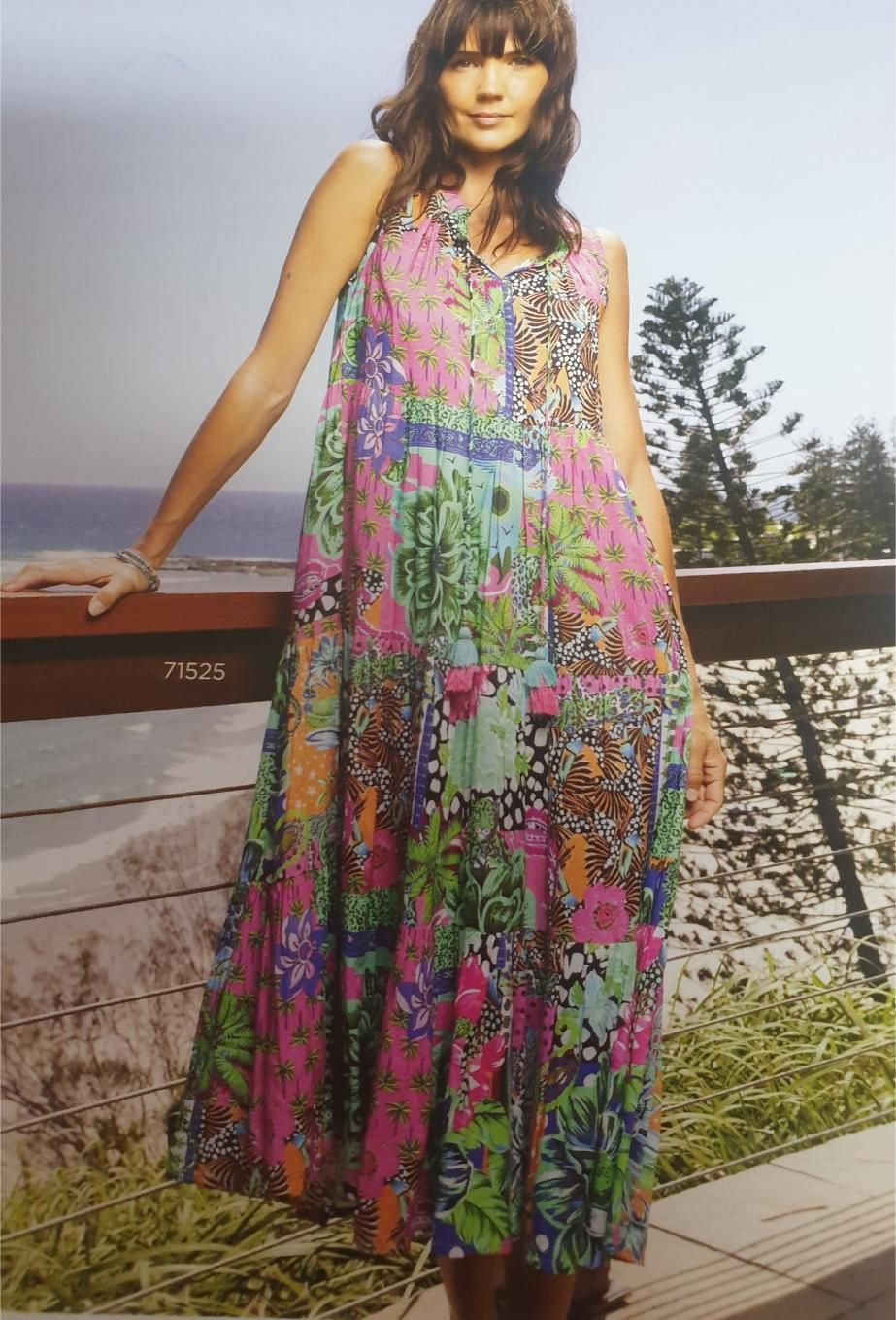 Woman in a brightly patterned maxi dress stands by a wooden railing overlooking the ocean.