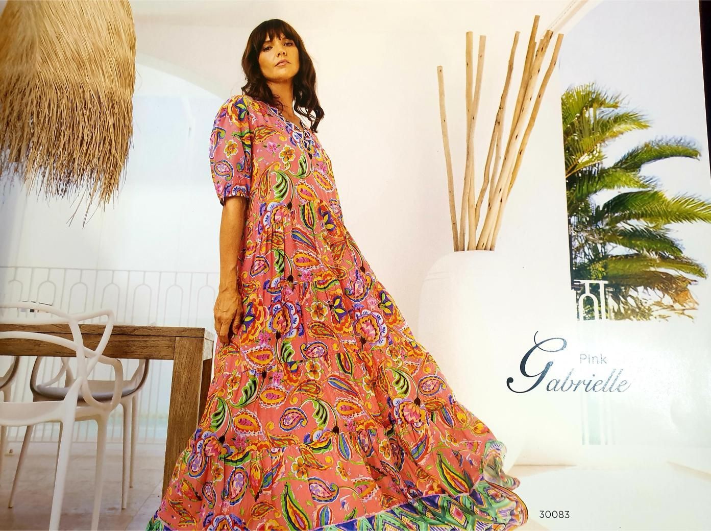 Woman in a flowy paisley dress, standing indoors near a table and window, with 