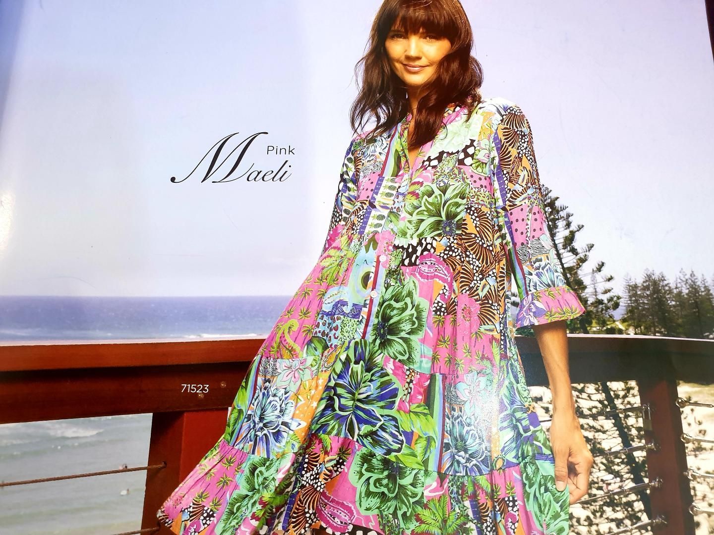Woman in colorful floral dress smiles on a deck overlooking the ocean.