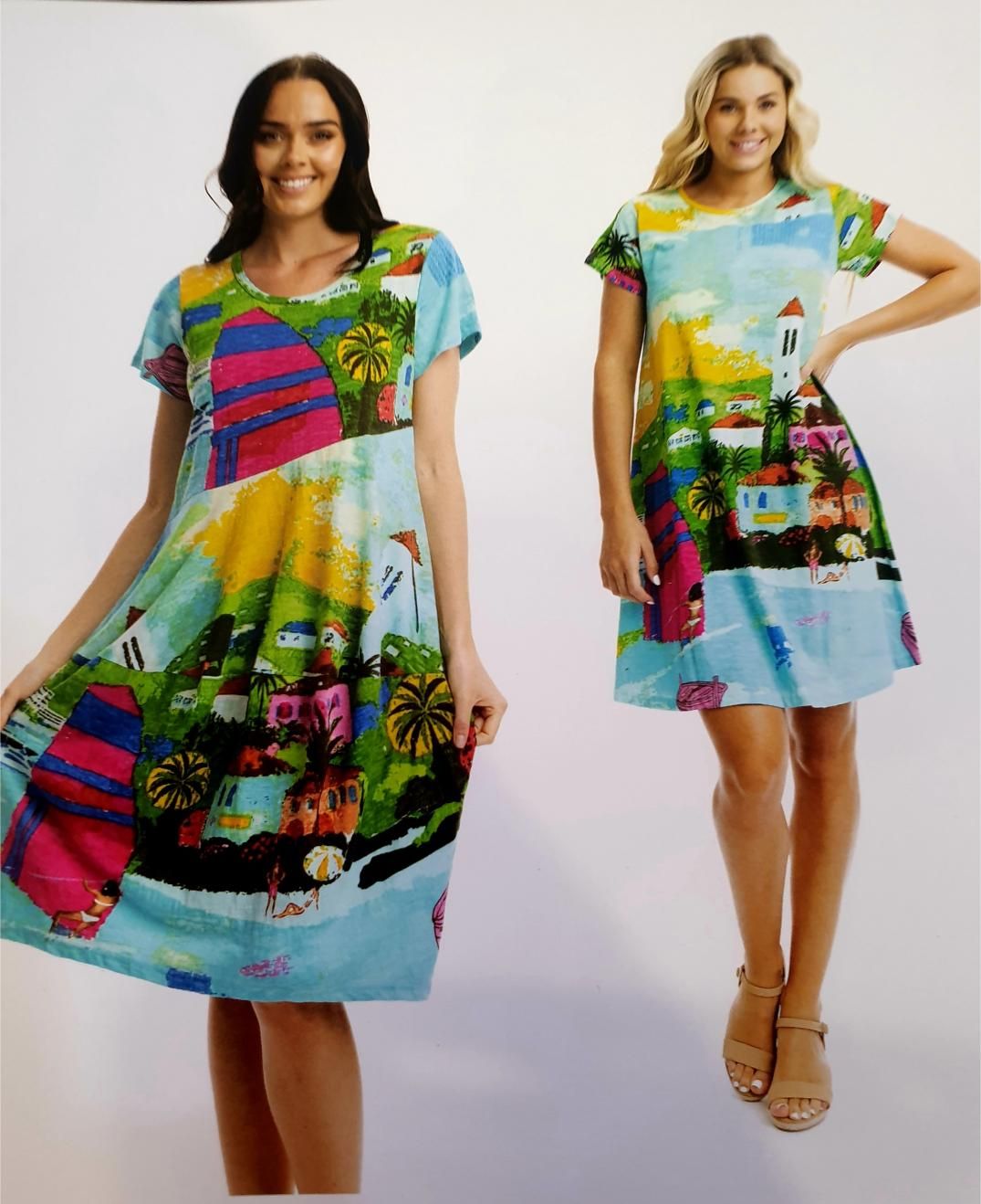 Two women wearing short-sleeved dresses with a colorful coastal scene print. One smiles, holding her dress out. The other poses with a hand on hip.