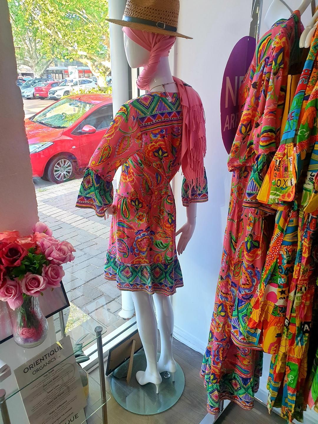 Mannequin in a pink patterned dress; shop display with colorful clothing. Window view of cars and trees.