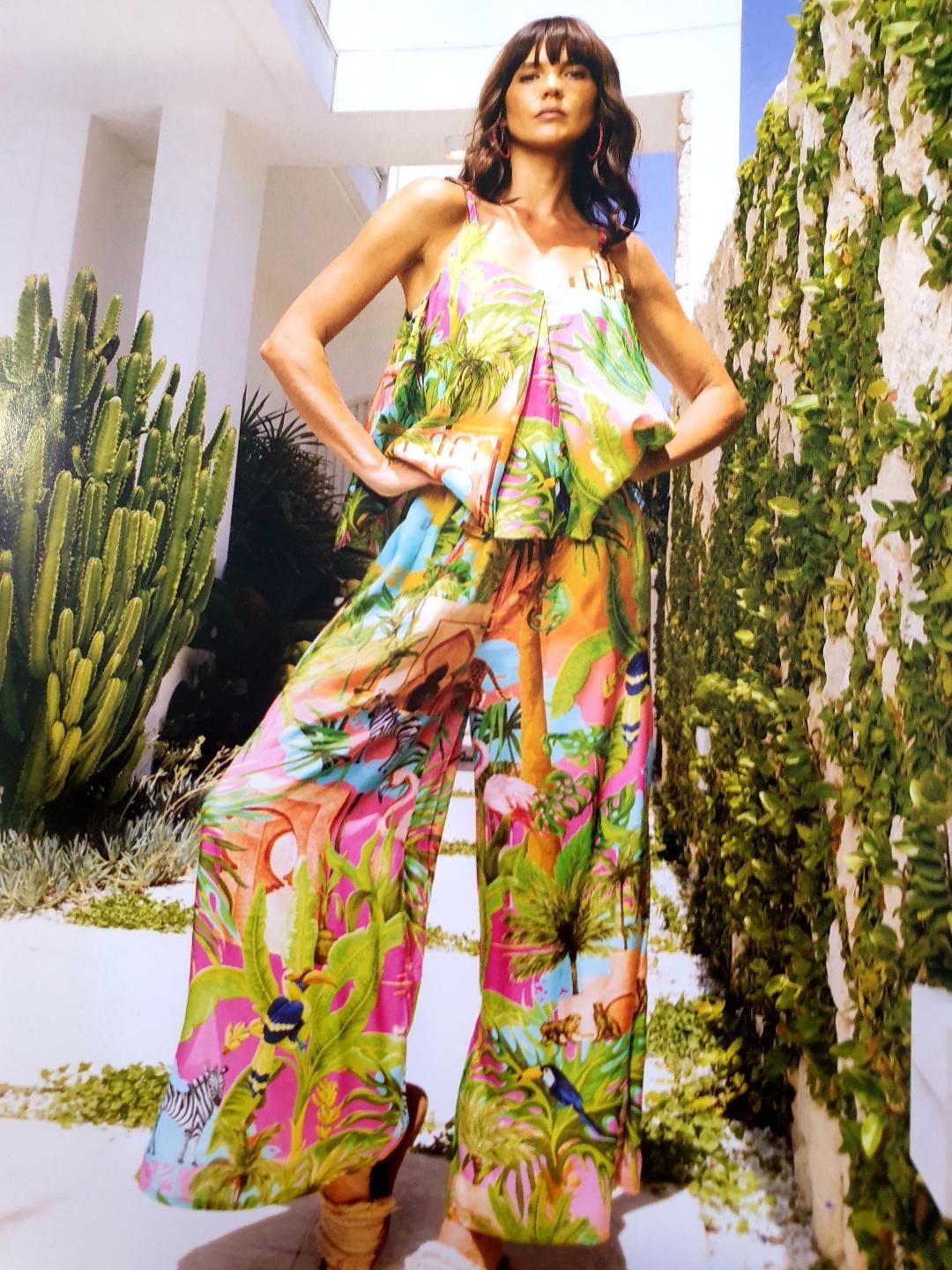 Woman in colorful tropical print jumpsuit, posing outdoors with hands on hips, bright setting.