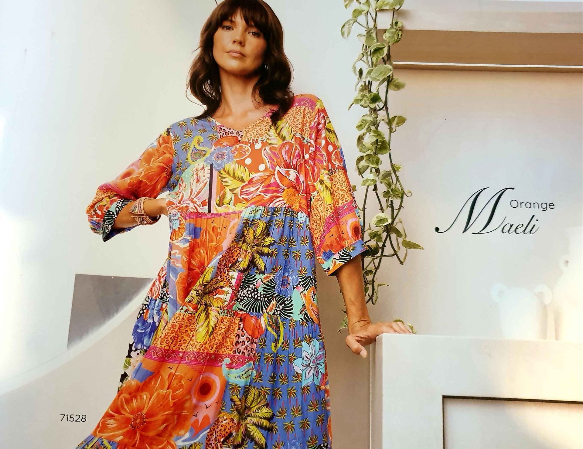 Woman in a colorful patterned dress leans on a white surface; vines in the background.