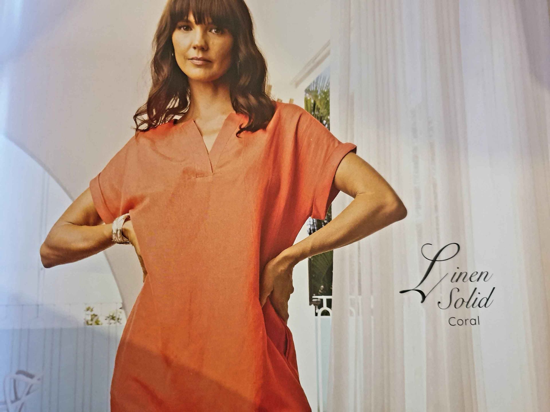 Woman in coral linen dress, posing with hands on hips, near curtains.