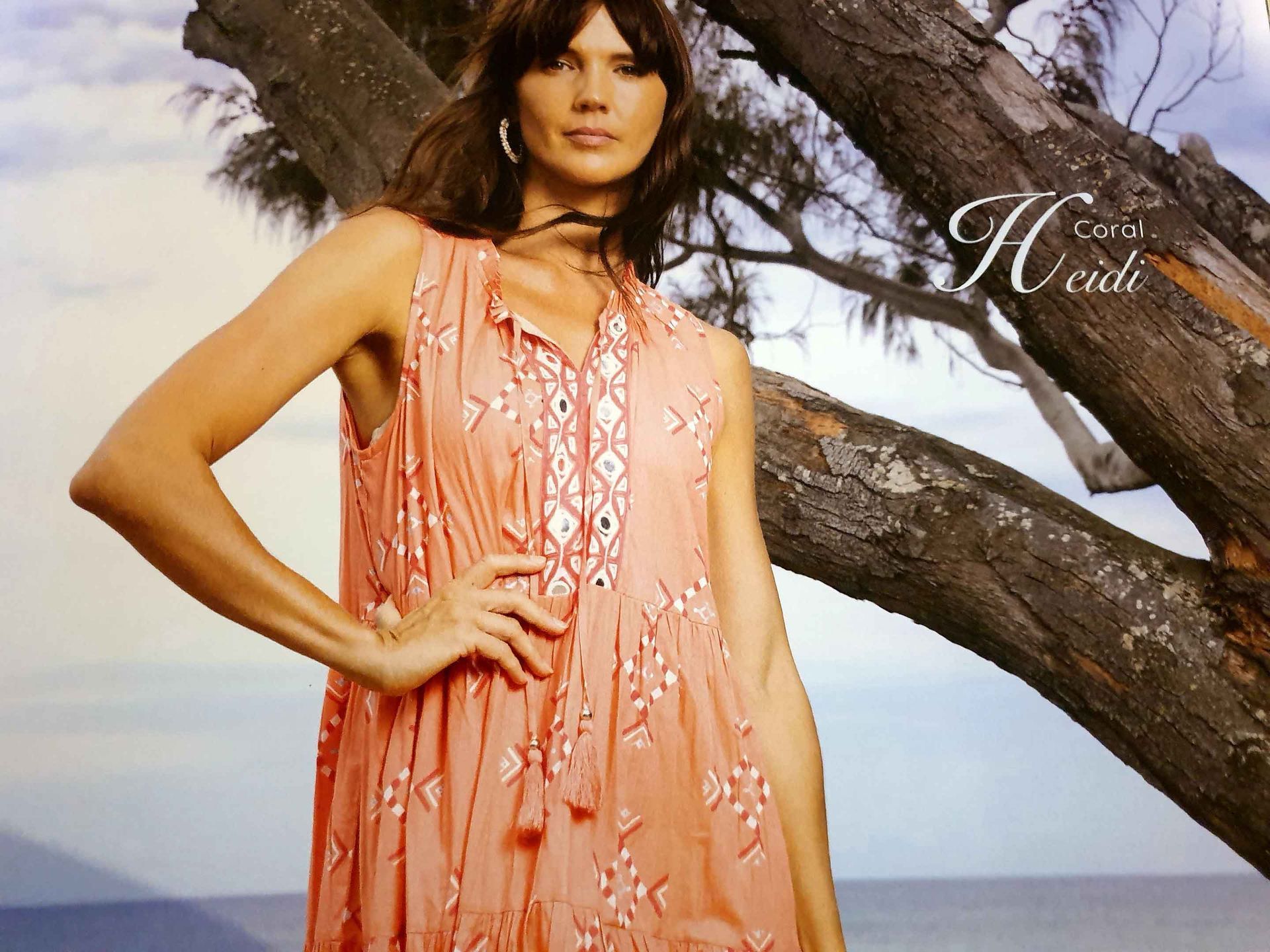 Woman in peach dress, hand on hip, by a tree, ocean background. 