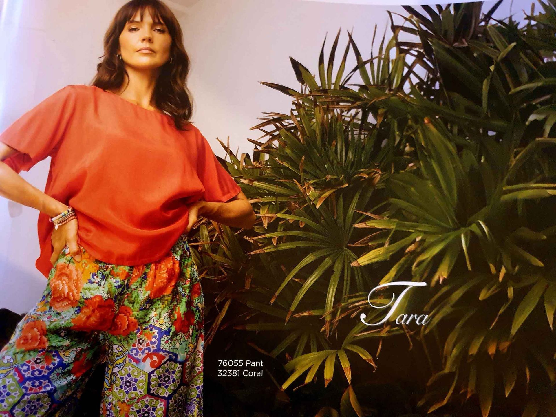 Woman wearing orange top and patterned pants, posing near tropical plants.