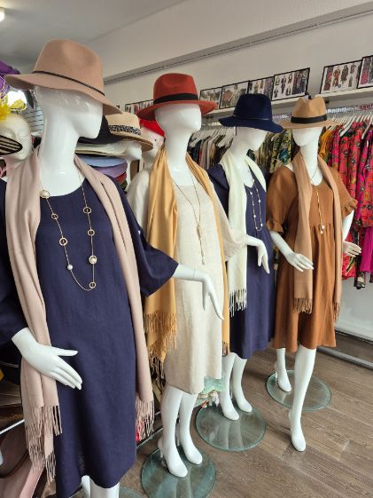 Four mannequins wearing dresses and hats in a boutique setting. Beige, rust, navy, and brown outfits are displayed.