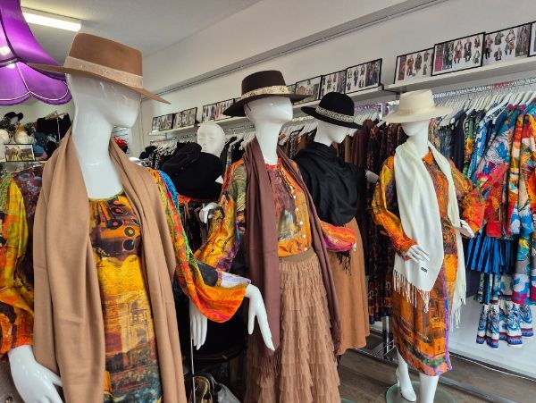 Mannequins in a shop wearing colorful patterned clothing, hats, and shawls. Clothes are on racks in the background.