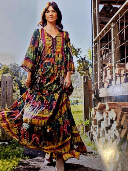 Woman in patterned maxi dress, walking outdoors near a wooden fence and stacked logs.