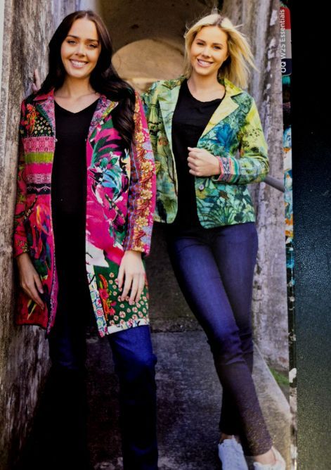 Two women in colorful jackets and jeans pose near a stone wall.