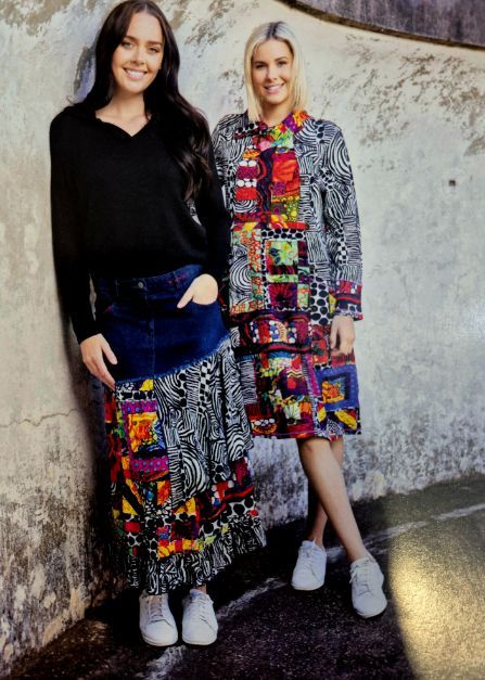 Two women in colorful outfits posing against a textured wall. One wears a black top and patterned denim skirt. The other wears a patterned coat dress.