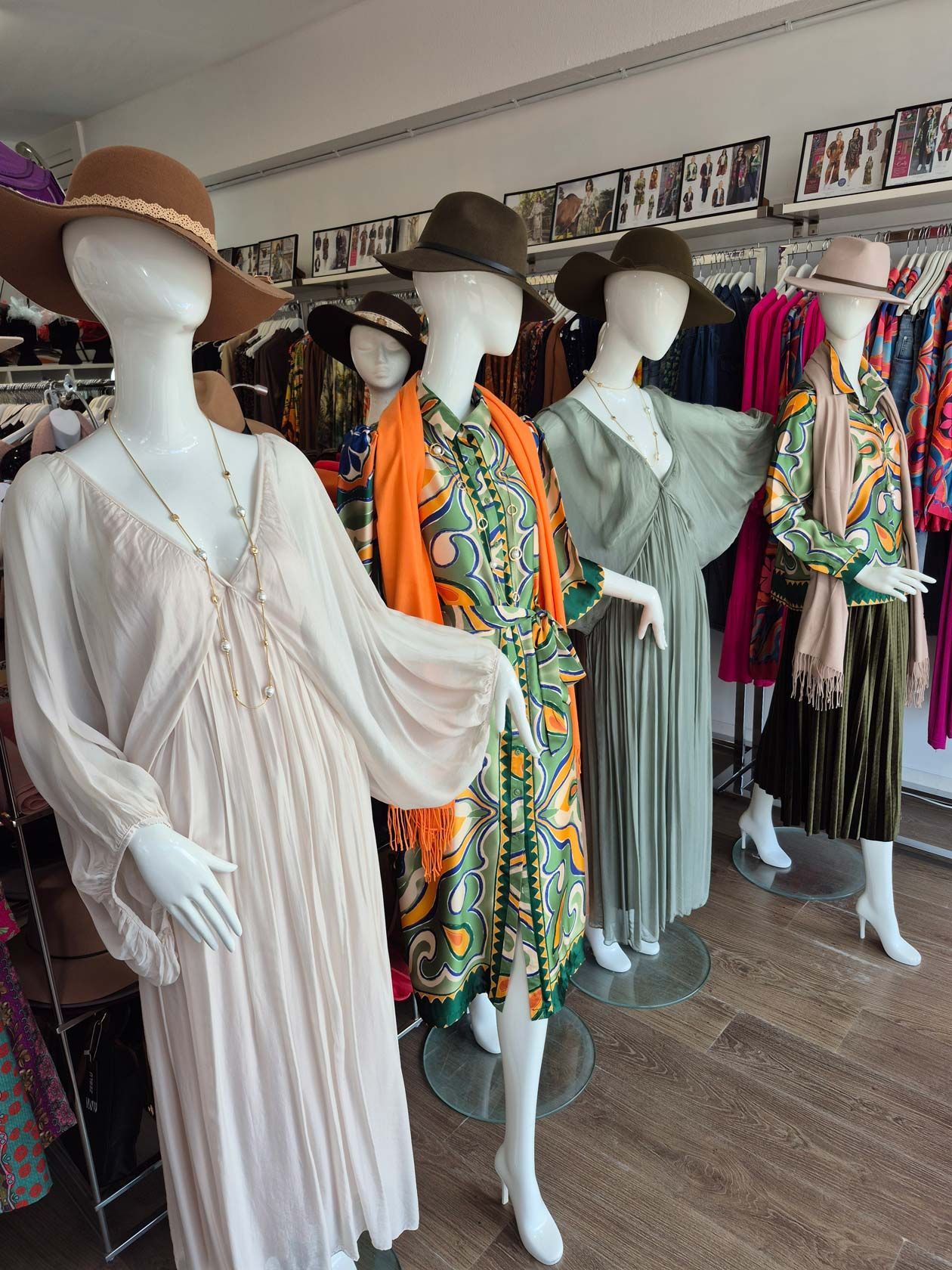 Five mannequins in a clothing store wearing hats, dresses, and a scarf. Beige, green, orange, and olive garments are displayed.