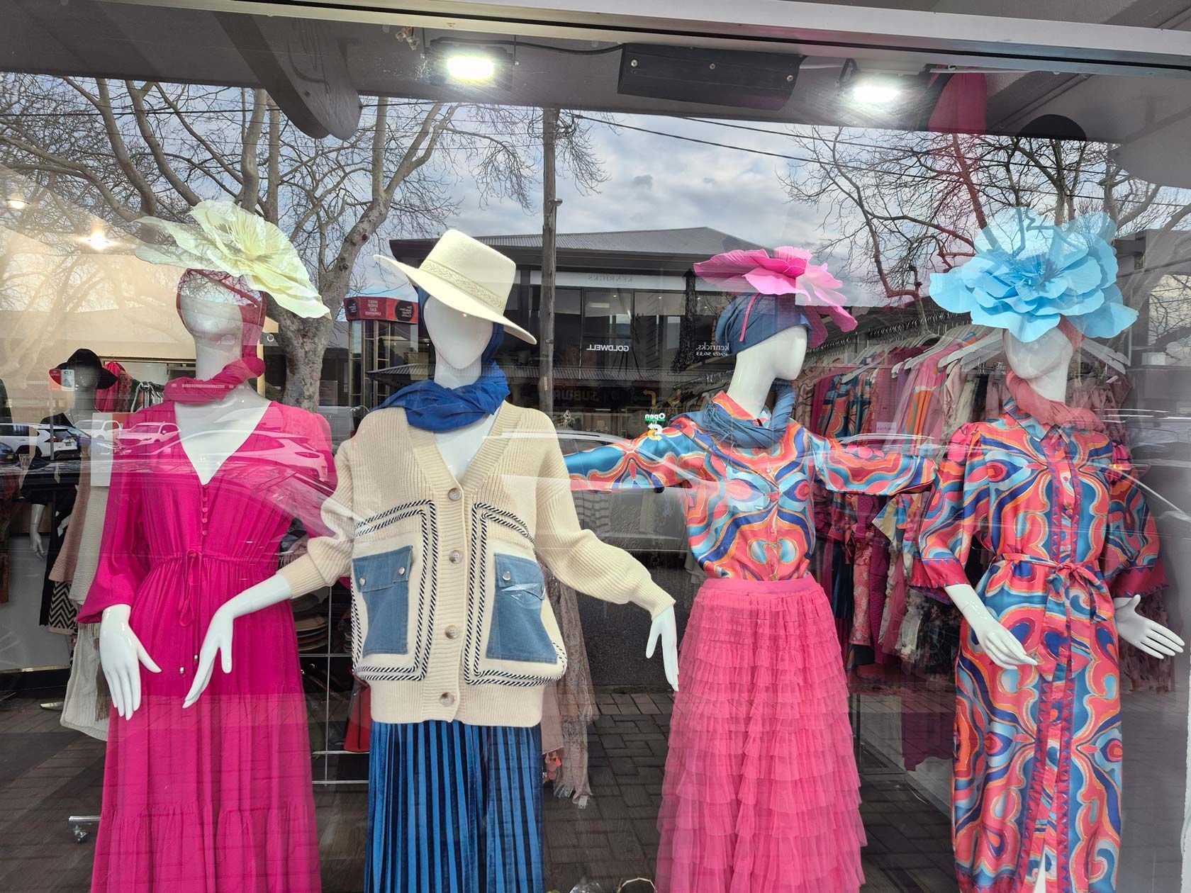 Four mannequins in a shop window display bright clothing and decorative hats.