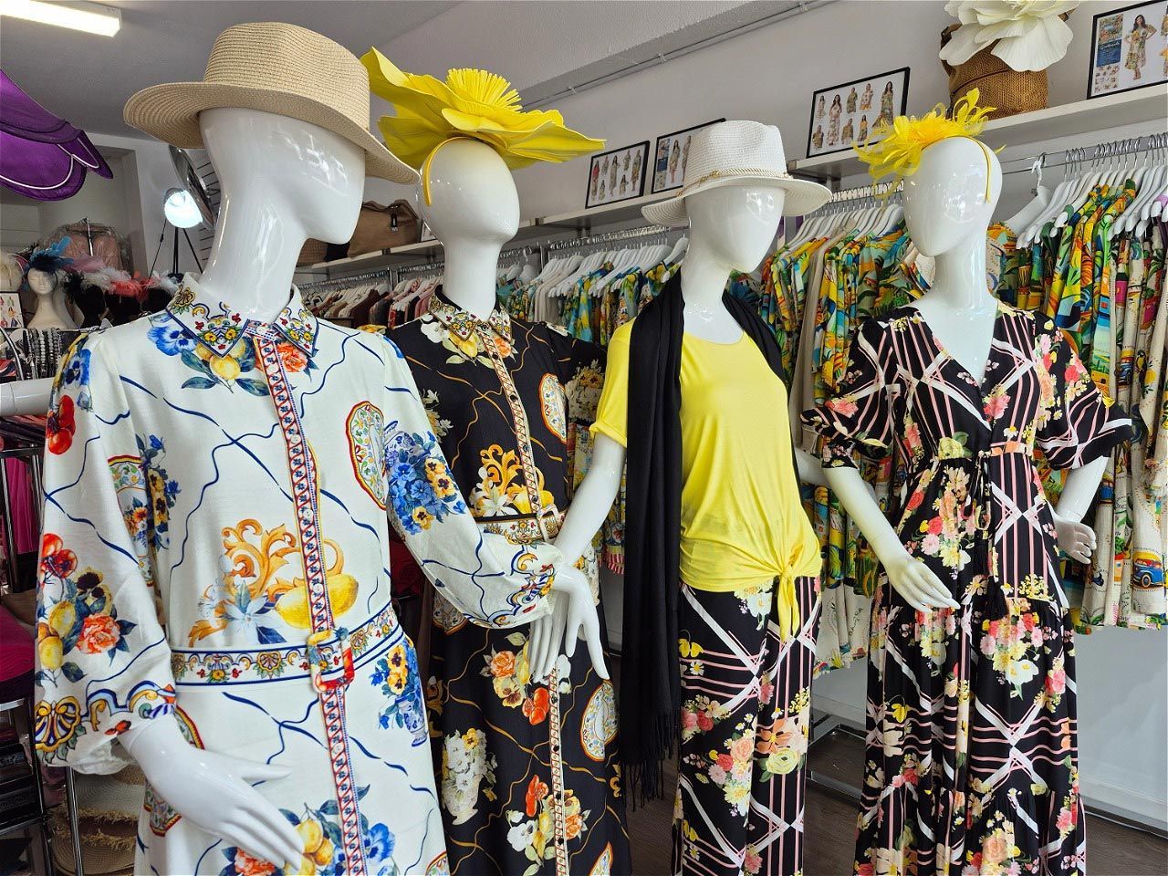 Four mannequins in a clothing store wearing floral dresses and hats; store with clothing racks.