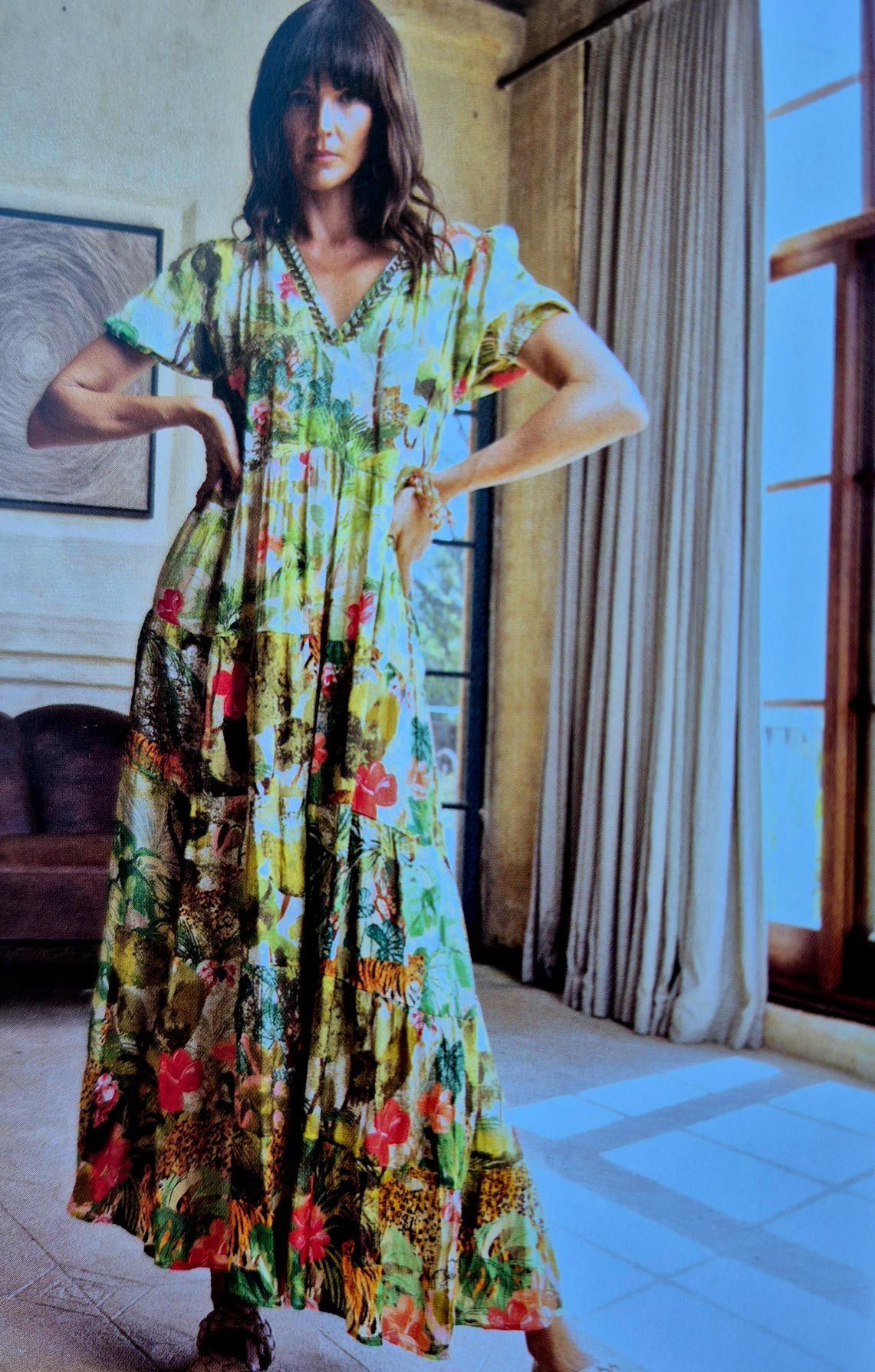 Woman in floral maxi dress, posing indoors by a window, hands on hips.